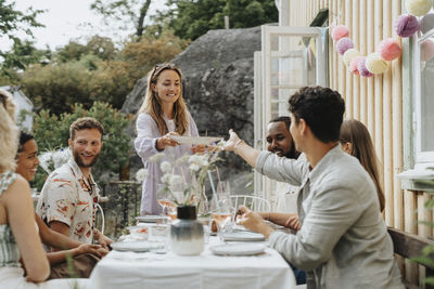 Smiling woman passing plate to male friend sitting at table during dinner party outside cafe