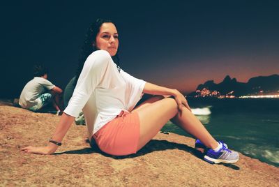 Side view of beautiful woman sitting on rocky shore at night