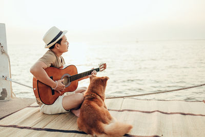 Side view of man playing guitar while sitting with dog at pier against sea