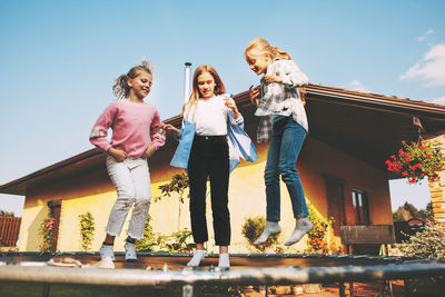 Full length of girls jumping on trampoline against house and sky