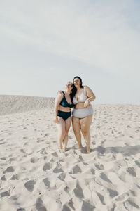 Glad curvy female friends in swimwear stand on sandy seashore and enjoying summer vacation together