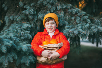 A cute boy in a bright orange jacket and a yellow hat holds a large package with oranges in his hand