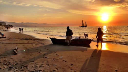 Silhouette people on beach by nautical vessel against sky during sunset