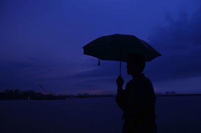 Rear view of man with umbrella on beach against sky during sunset