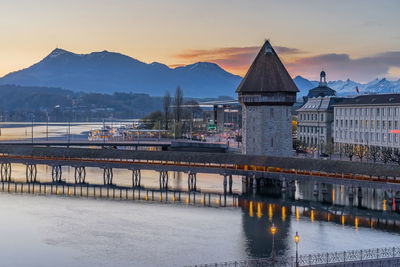 Bridge over river by buildings against sky during sunset