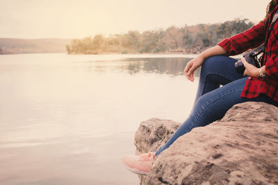 Young woman sitting on rock by lake against clear sky