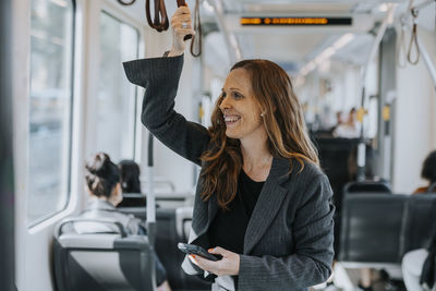 Happy female passenger holding smart phone while standing in train