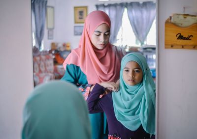 Mother helping daughter in getting dressed by mirror at home