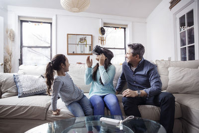 Father and daughter looking at girl wearing virtual reality simulator while sitting on sofa