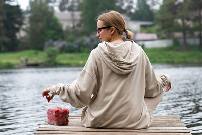 Young woman eating berry fruit strawberries in plastic container, resting on a river wooden pier