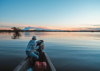 Man sitting on lake against sky during sunset