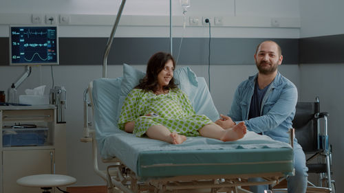 Smiling man sitting by pregnant wife in hospital ward