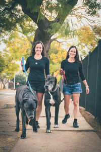 Portrait of smiling sisters with dog walking on footpath against trees