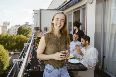 Portrait of smiling woman holding drink glass while celebrating dinner party with friends in balcony