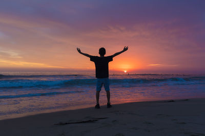 Rear view of man with arms outstretched standing against sea during sunset