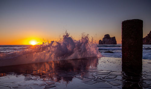 Sea waves splashing on shore against sky during sunset