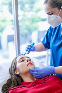 Esthetician in uniform and face mask injecting collagen into forehead of female client near window in office of beauty clinic.