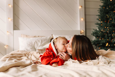 Happy mom and daughter baby in red hugging lying on the bed in the room with a christmas tree