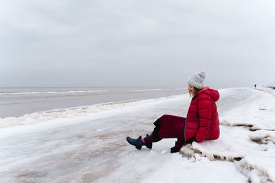A cheerful woman in a red jacket and a warm dress sits on the shore of the winter sea