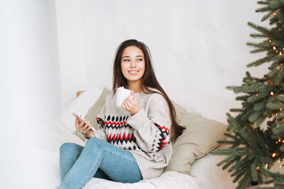 Young asian woman with dark long hair using mobile smartphone in room with christmas tree at home