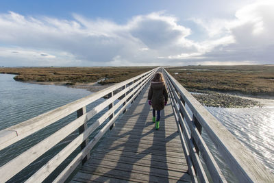 Rear view of girl walking on railing by river against sky