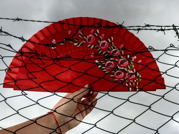 Cropped hand holding red hand fan against sky