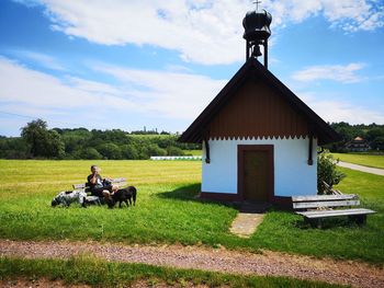 Man with dogs sitting on grass by house against sky