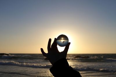Cropped hand holding crystal ball at beach against sky during sunset