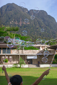 Rear view of man playing bubble with arms raised standing against mountain