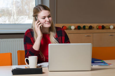 Businesswoman talking on mobile phone while using laptop in office
