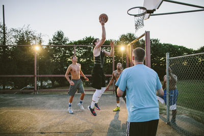 Friends looking at man dunking while practicing basketball in court during sunset