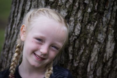 Portrait of cute smiling girl with blond hair standing against tree trunk