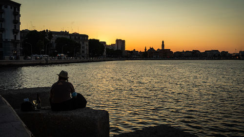 Rear view of man sitting on river against buildings at sunset