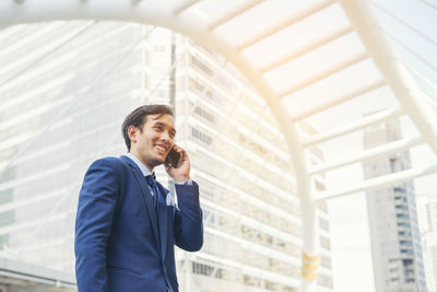 Smiling young businessman using mobile phone against buildings in city