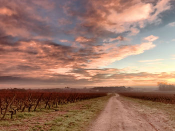 Dirt road amidst field against sky during sunset