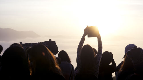 Rear view of woman taking selfie with friends against sky during sunset
