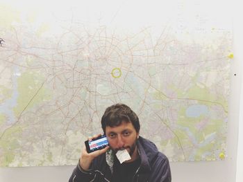 Portrait of man using phone while carrying paper in mouth by map on wall