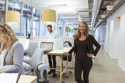 Portrait of smiling businesswoman standing at desk while colleagues working in office