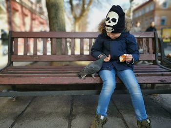 Boy feeding pigeon while sitting on bench in city