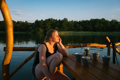 Young woman with eyes closed sitting by lake during sunset