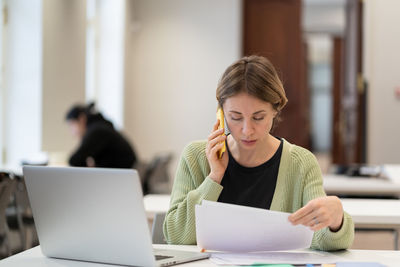 Middle-aged woman busy with documents talking on cellphone working in campus open space. education.