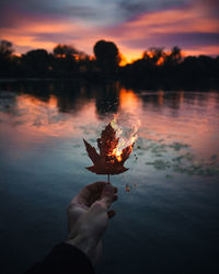 Cropped hand holding burning autumn leaf against lake during sunset