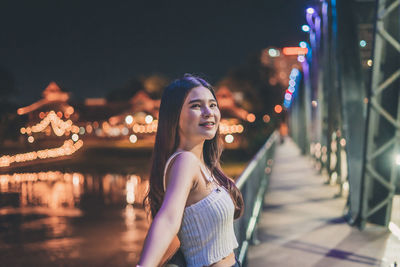 Portrait of smiling young woman standing on bridge over river at night