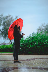 Woman with a mask holding a red umbrella looks up while raining