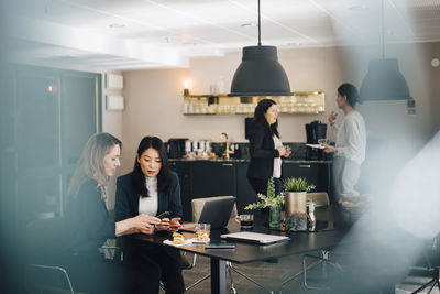 Businesswomen using smart phones at conference table while colleagues standing in background