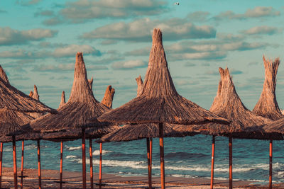 Thatched roofs at beach against cloudy sky during sunset