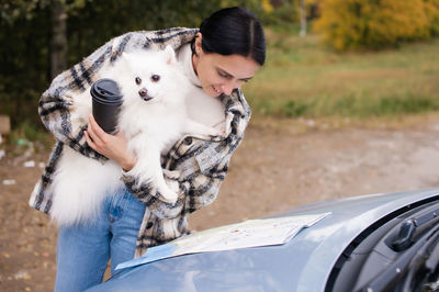 The girl is standing by the car holding a pomeranian in her hands looking at the road map on 