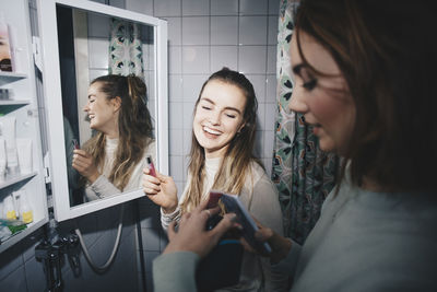 Happy female friends with mobile phone and lip gloss at dorm bathroom