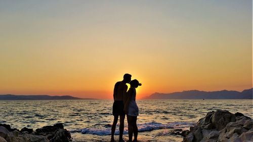 Couple kissing while standing on shore at beach against sky during sunset