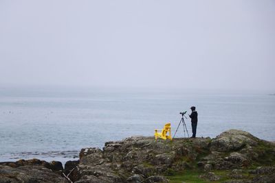 Man photographing while standing on rock by sea against sky